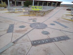 Photograph of the Four Corners Monument, the only point in the United States where four states meet: Arizona, Utah, Colorado, and New Mexico. The ground marker is set in a paved plaza with state names and official seals inlaid in metal. A yellow "Must Wear Mask" public health order sign is visible on a railing in the background, photographed during the COVID-19 pandemic. Photo by Mx. Granger, CC0, via Wikimedia Commons.