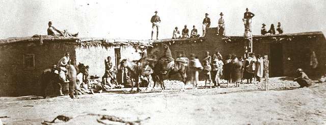 Historic photograph of Navajo (Diné) captives at Fort Sumner, New Mexico, circa 1860s. A large adobe structure stretches across the background, with people standing on its roof. In the foreground, a crowd of people and a rider on horseback are gathered in an open dirt yard.